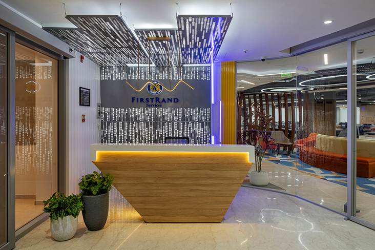Corporate reception lobby interior design by Trimit Rachana featuring FirstRand branding, custom angular wooden desk, decorative ceiling feature, and modern office lighting.